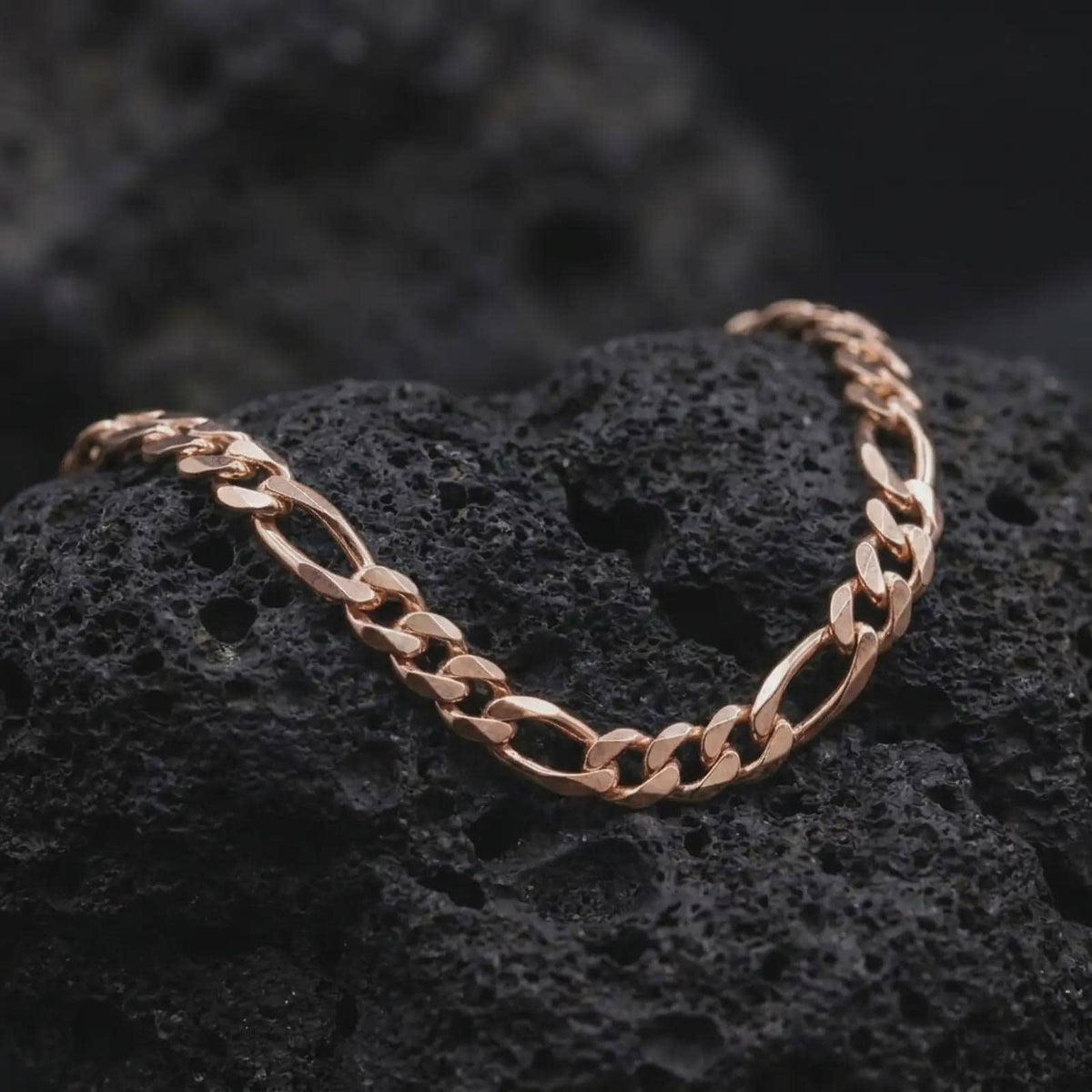 The rose gold plated 7mm Figaro chain necklace rests on black volcanic rock, emphasizing its articulated Italian link construction and bold masculine profile in a detailed macro image.