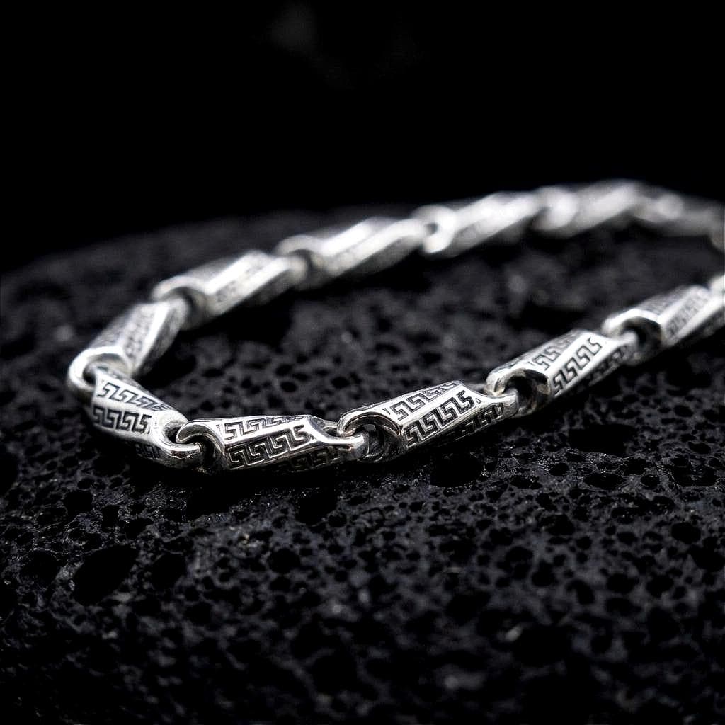 The polished silver Greek meander necklace is displayed across textured black volcanic rock, highlighting the repeating ancient Greek key pattern and articulated link construction in a detailed macro photograph.