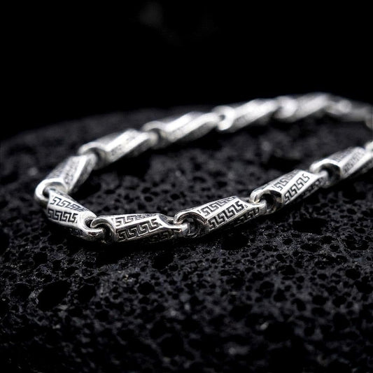 The polished silver Greek meander necklace is displayed across textured black volcanic rock, highlighting the repeating ancient Greek key pattern and articulated link construction in a detailed macro photograph.