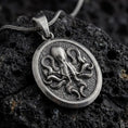 Load image into Gallery viewer, An oxidized sterling silver octopus pendant rests on dark volcanic rock, showing raised tentacles, detailed suction cups, and a textured round medallion background in a macro studio close-up.
