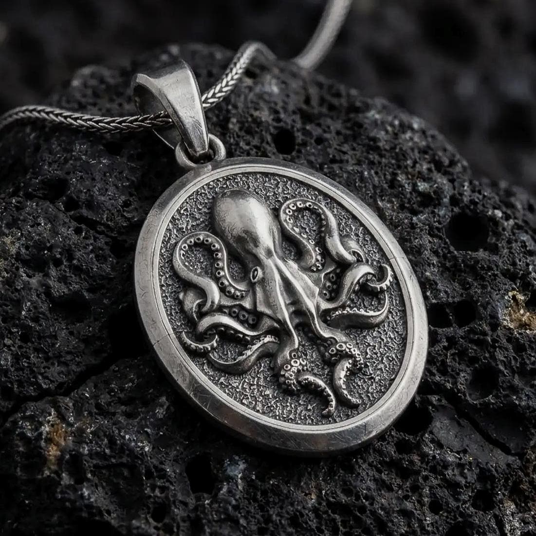An oxidized sterling silver octopus pendant rests on dark volcanic rock, showing raised tentacles, detailed suction cups, and a textured round medallion background in a macro studio close-up.