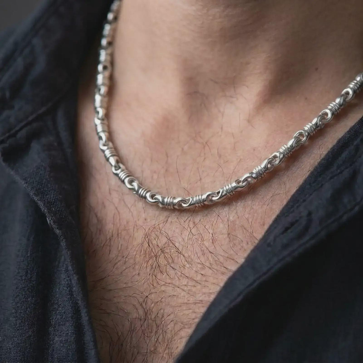 The polished silver cylinder link necklace is worn on a man’s chest, showing the repeating barrel sections and articulated chain movement in a premium lifestyle photograph.