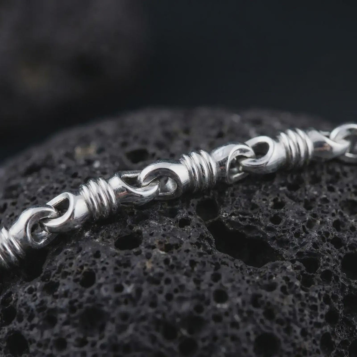 The polished silver barrel chain necklace is displayed on textured black volcanic rock, highlighting the raised ridges, smooth link openings, and articulated sterling silver construction in a premium macro photograph.