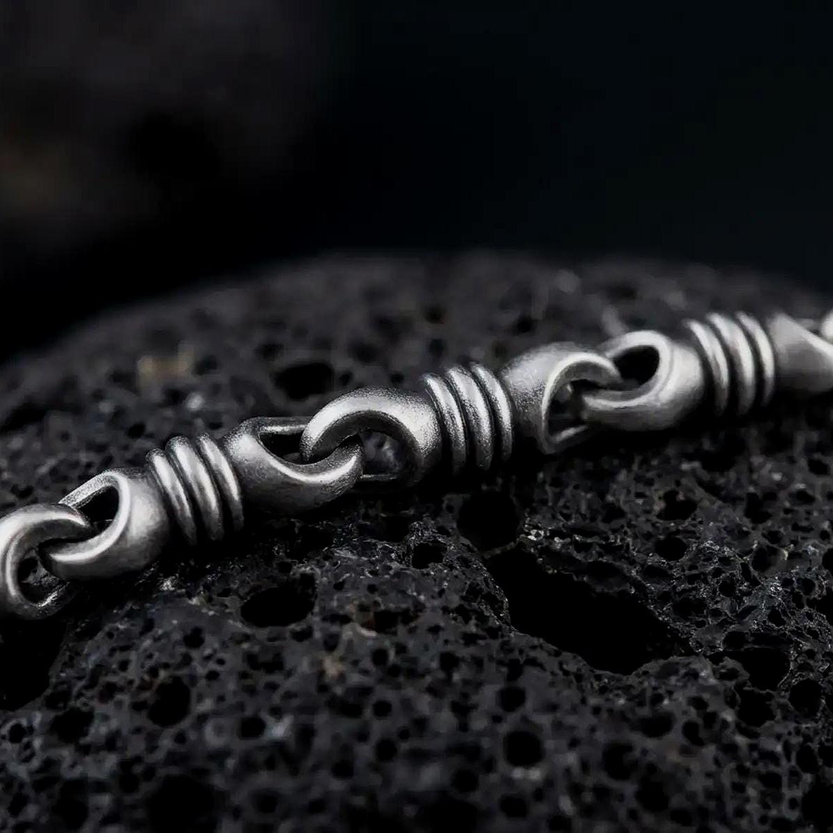 A ridged barrel chain necklace with an oxidized finish rests on dark volcanic rock, showing rounded connector links and sculpted cylinder sections in a macro studio close-up.