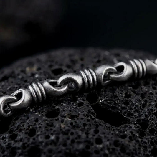 A ridged barrel chain necklace with an oxidized finish rests on dark volcanic rock, showing rounded connector links and sculpted cylinder sections in a macro studio close-up.