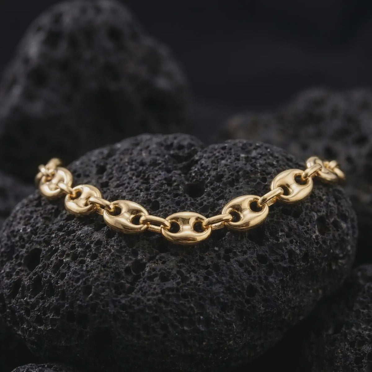 A gold plated 11mm anchor link necklace is displayed on rugged volcanic rock, featuring bold oval mariner links divided by distinctive central bars in a luxury macro studio close-up.