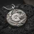 Load image into Gallery viewer, The polished silver dragon and phoenix pendant is displayed on textured black volcanic rock, highlighting the sculpted mythical creatures surrounding a geometric Yin Yang centerpiece in a premium macro photograph.
