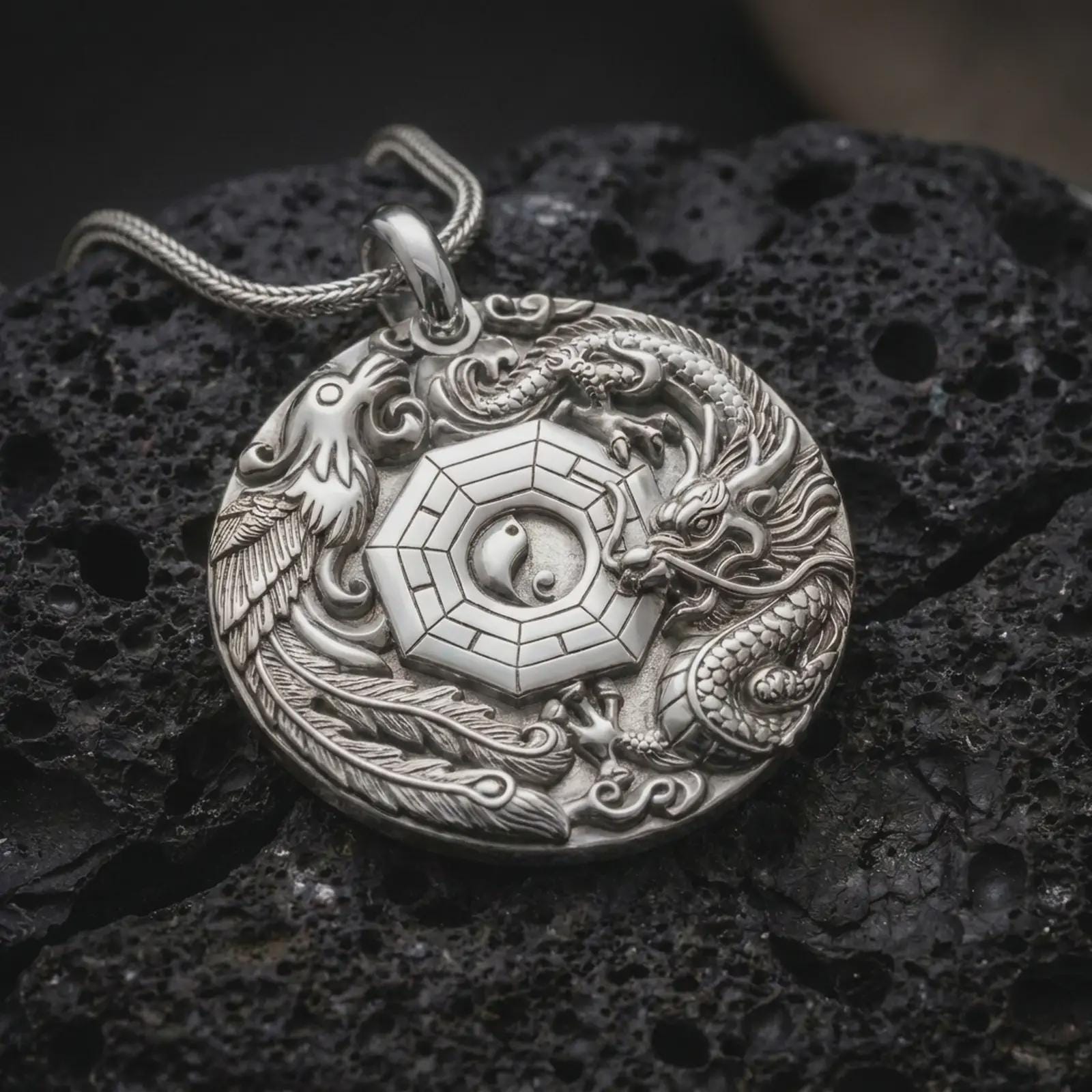 The polished silver dragon and phoenix pendant is displayed on textured black volcanic rock, highlighting the sculpted mythical creatures surrounding a geometric Yin Yang centerpiece in a premium macro photograph.