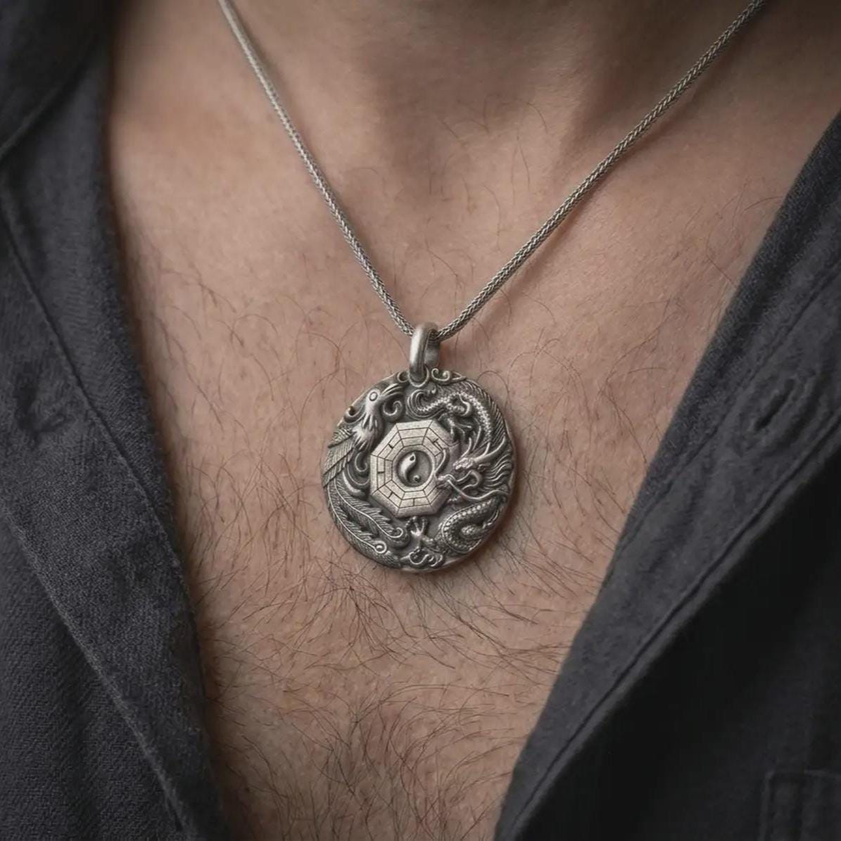 An oxidized dragon and phoenix pendant is worn on a man’s neck, displaying the dark recessed details, central Yin Yang symbol, Bagua geometry, and mythical creature relief in a masculine lifestyle close-up.