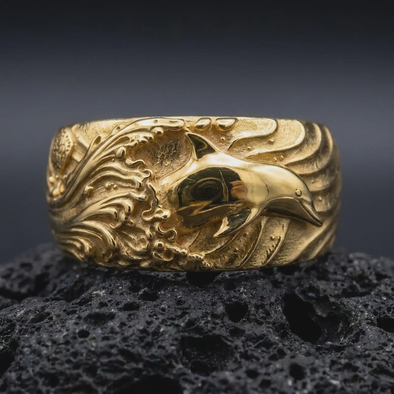 A gold plated dolphin ring rests on rugged volcanic rock, featuring a dimensional dolphin surrounded by flowing ocean waves and textured sea details in a luxury macro studio close-up.