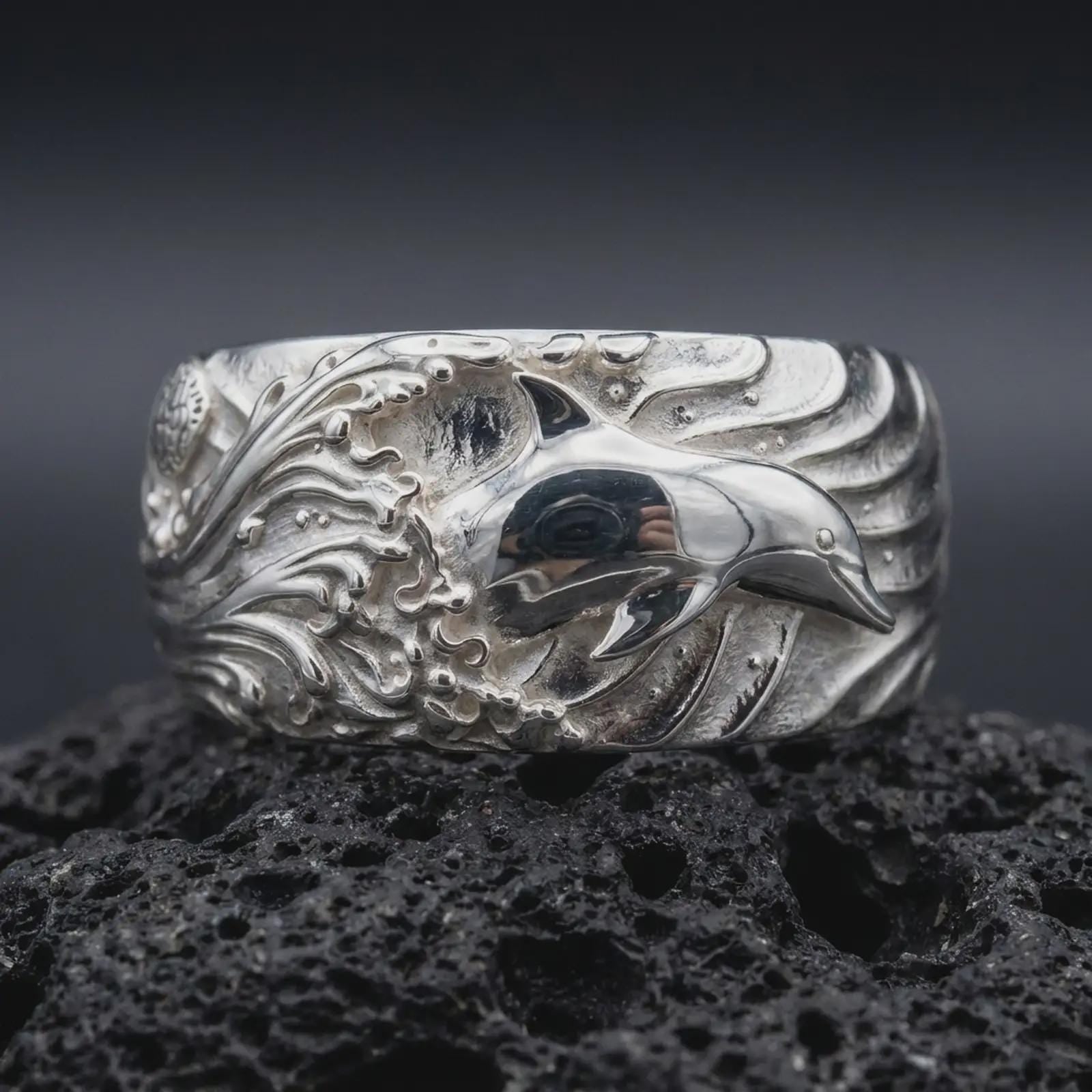 The polished silver dolphin band is displayed on textured black volcanic rock, highlighting the smooth marine figure, layered wave relief, and broad sculptural profile in a detailed macro photograph.