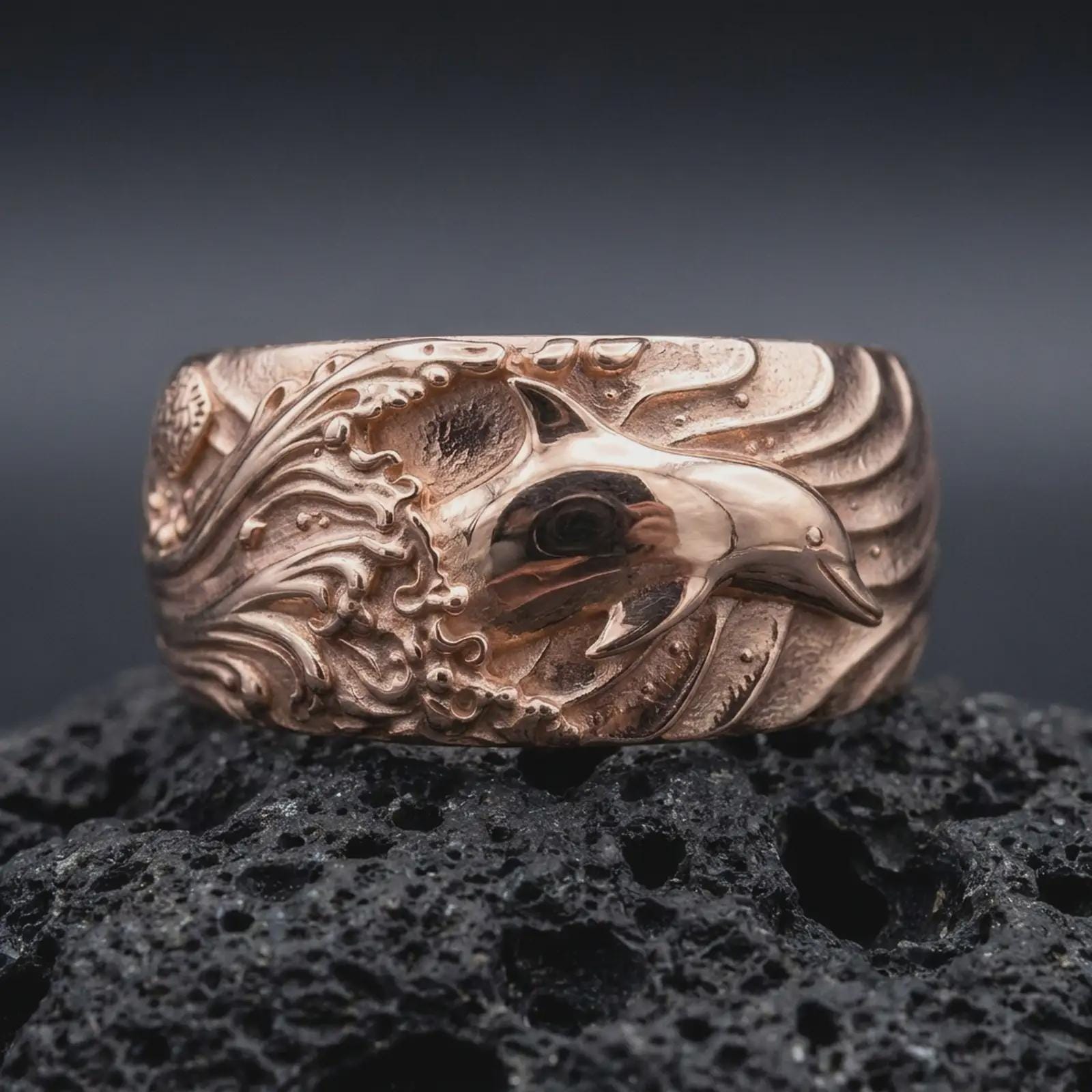The rose gold plated ocean wave ring is arranged on black volcanic rock, emphasizing the raised dolphin motif, curling wave patterns, and substantial unisex band in a refined macro image.
