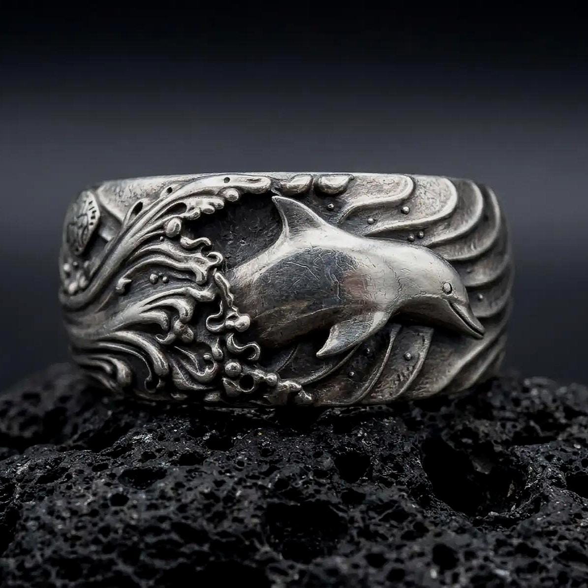 An oxidized sterling silver dolphin ring rests on dark volcanic rock, showing a raised dolphin swimming through deeply sculpted ocean waves in a premium macro studio close-up.