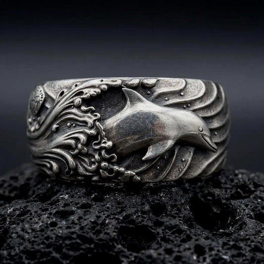 An oxidized sterling silver dolphin ring rests on dark volcanic rock, showing a raised dolphin swimming through deeply sculpted ocean waves in a premium macro studio close-up.