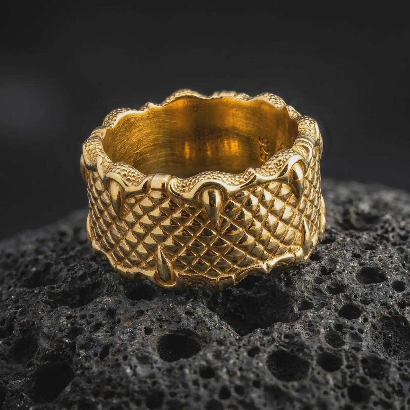 A gold plated dragon scale ring rests on rugged volcanic rock, featuring dimensional reptile scales and pointed claws wrapped around the upper and lower edges in a luxury macro studio close-up.