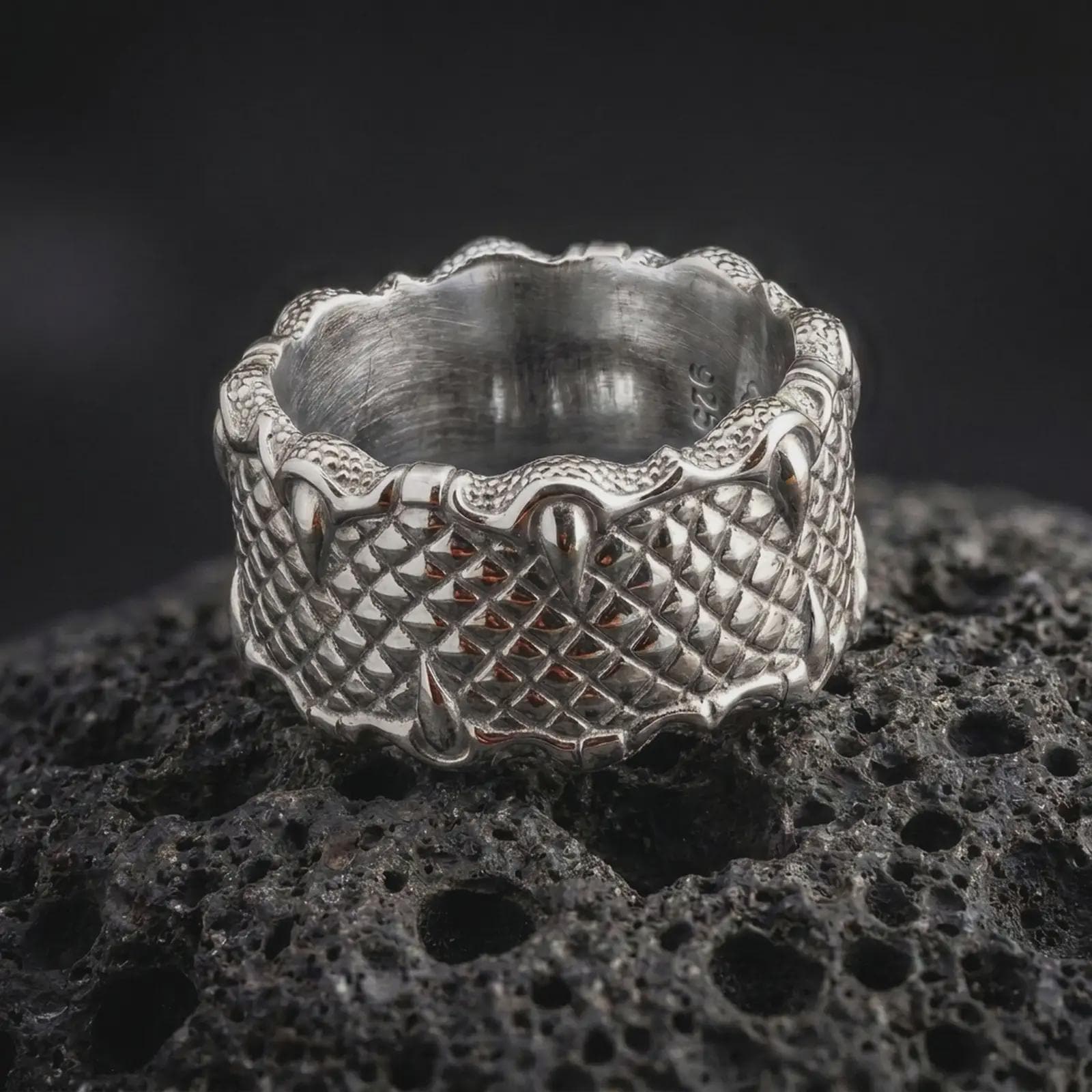 The polished silver dragon claw ring is displayed on textured black volcanic rock, highlighting the repeating scale pattern, sculpted talons, and substantial unisex band in a detailed macro photograph.