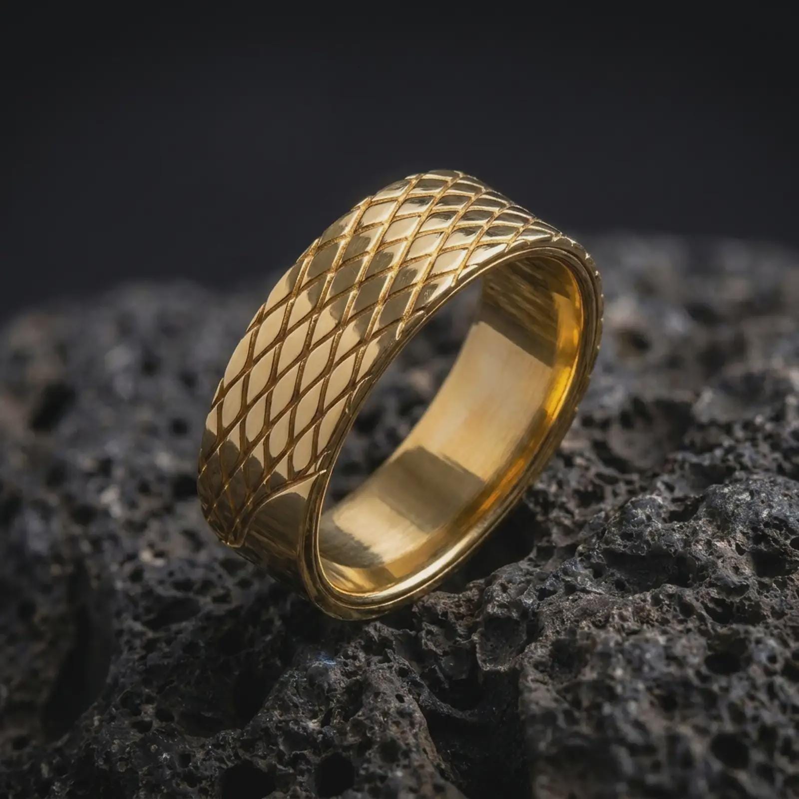 A gold plated crosshatch ring rests on rugged volcanic rock, featuring a repeating diagonal lattice pattern across its wide unisex band in a luxury macro studio close-up.