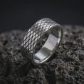 Load image into Gallery viewer, The polished silver geometric band is displayed upright on textured black volcanic rock, highlighting the intersecting engraved lines, raised diamond sections, and smooth inner surface in a detailed macro photograph.
