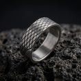 Load image into Gallery viewer, An oxidized sterling silver lattice ring stands on dark volcanic rock, showing its continuous diamond-shaped crosshatch pattern and recessed diagonal grooves in a premium macro studio close-up.
