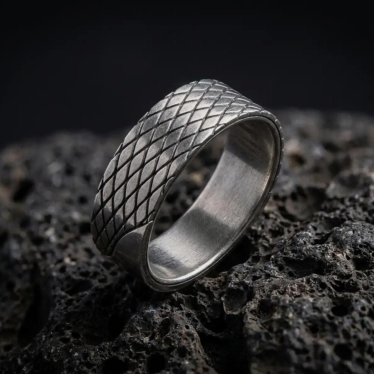 An oxidized sterling silver lattice ring stands on dark volcanic rock, showing its continuous diamond-shaped crosshatch pattern and recessed diagonal grooves in a premium macro studio close-up.