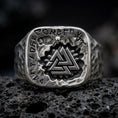 Load image into Gallery viewer, An oxidized sterling silver Valknut ring rests on dark volcanic rock, showing the raised interlocking triangle symbol, engraved Norse runes, skull accent, and rugged square signet face in a macro studio close-up.
