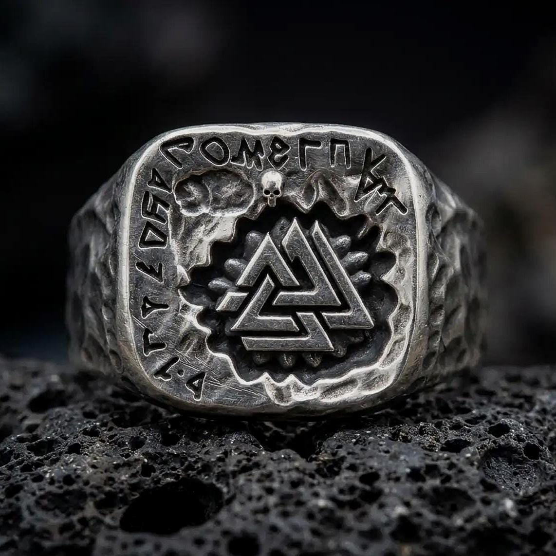 An oxidized sterling silver Valknut ring rests on dark volcanic rock, showing the raised interlocking triangle symbol, engraved Norse runes, skull accent, and rugged square signet face in a macro studio close-up.