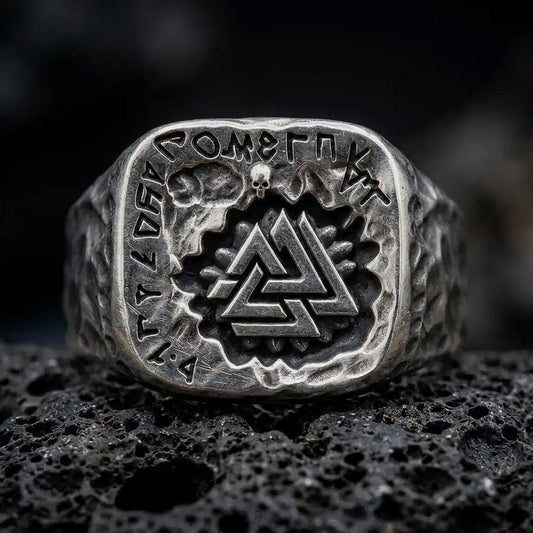 An oxidized sterling silver Valknut ring rests on dark volcanic rock, showing the raised interlocking triangle symbol, engraved Norse runes, skull accent, and rugged square signet face in a macro studio close-up.