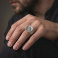 Load image into Gallery viewer, The polished silver Viking rune ring is worn on a man’s finger, with the square signet face, central Valknut emblem, engraved border, and sculpted shoulders clearly visible in a premium lifestyle photograph.
