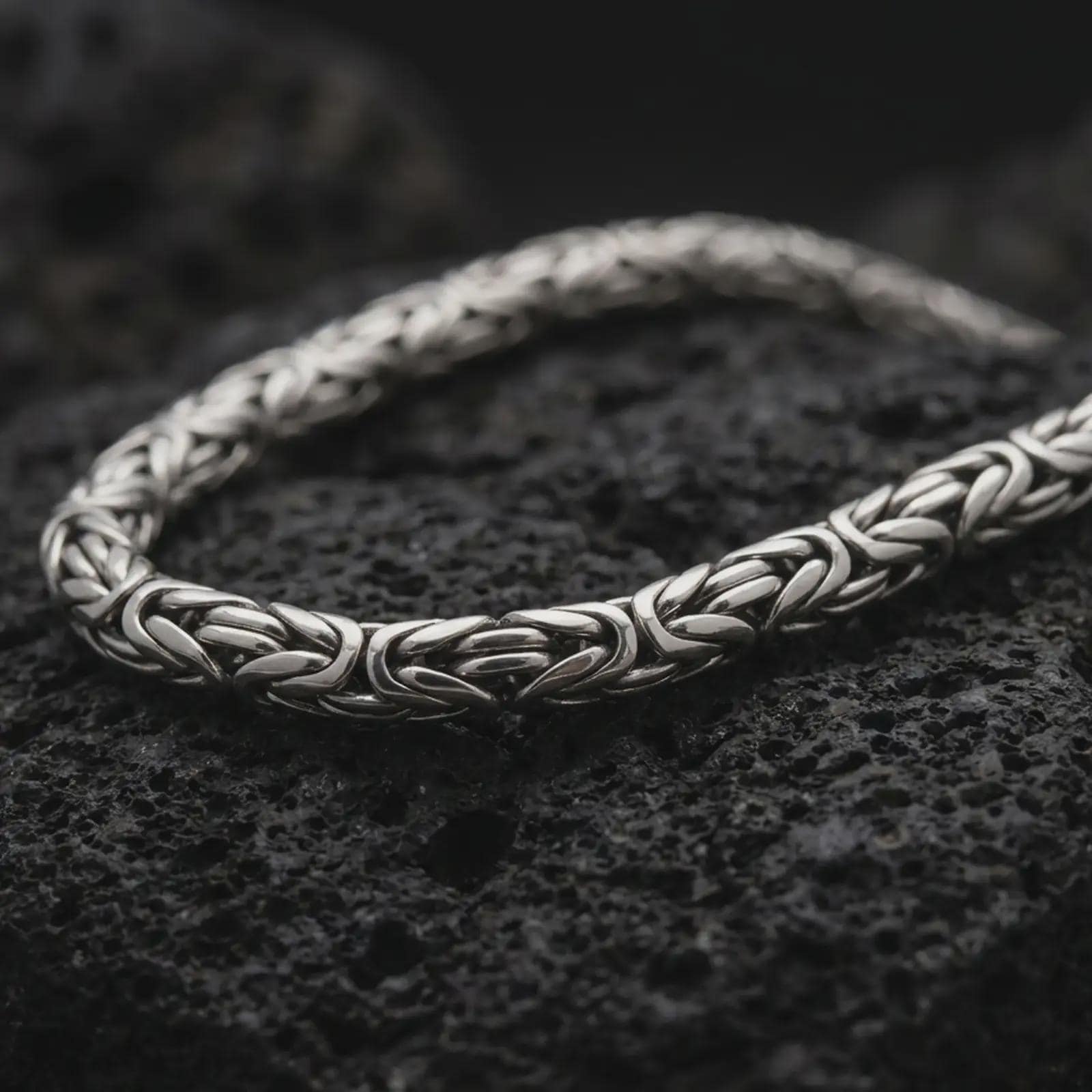 A 6mm Byzantine chain necklace with an oxidized finish rests on dark volcanic rock, showing its rounded interlocking links and woven sterling silver structure in a macro studio close-up.
