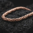Load image into Gallery viewer, The rose gold plated 6mm Byzantine necklace is arranged on black volcanic rock, emphasizing its rounded woven link pattern, flexible chain structure, and refined statement design in a detailed macro image.
