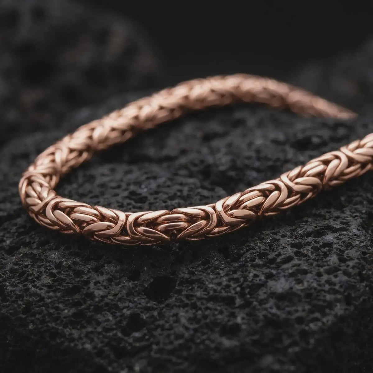 The rose gold plated 6mm Byzantine necklace is arranged on black volcanic rock, emphasizing its rounded woven link pattern, flexible chain structure, and refined statement design in a detailed macro image.