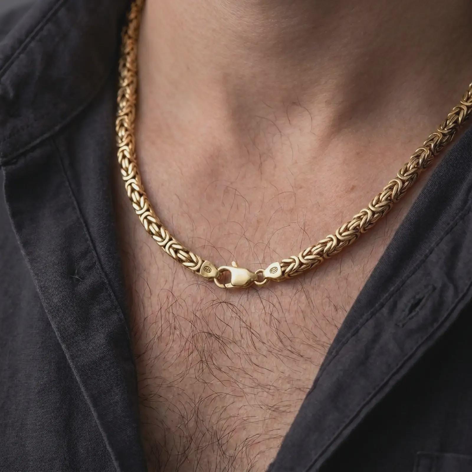 A gold plated 6mm Byzantine chain necklace is worn on a man’s neck with dark clothing, highlighting the woven link construction and strong statement profile in a refined close-up.