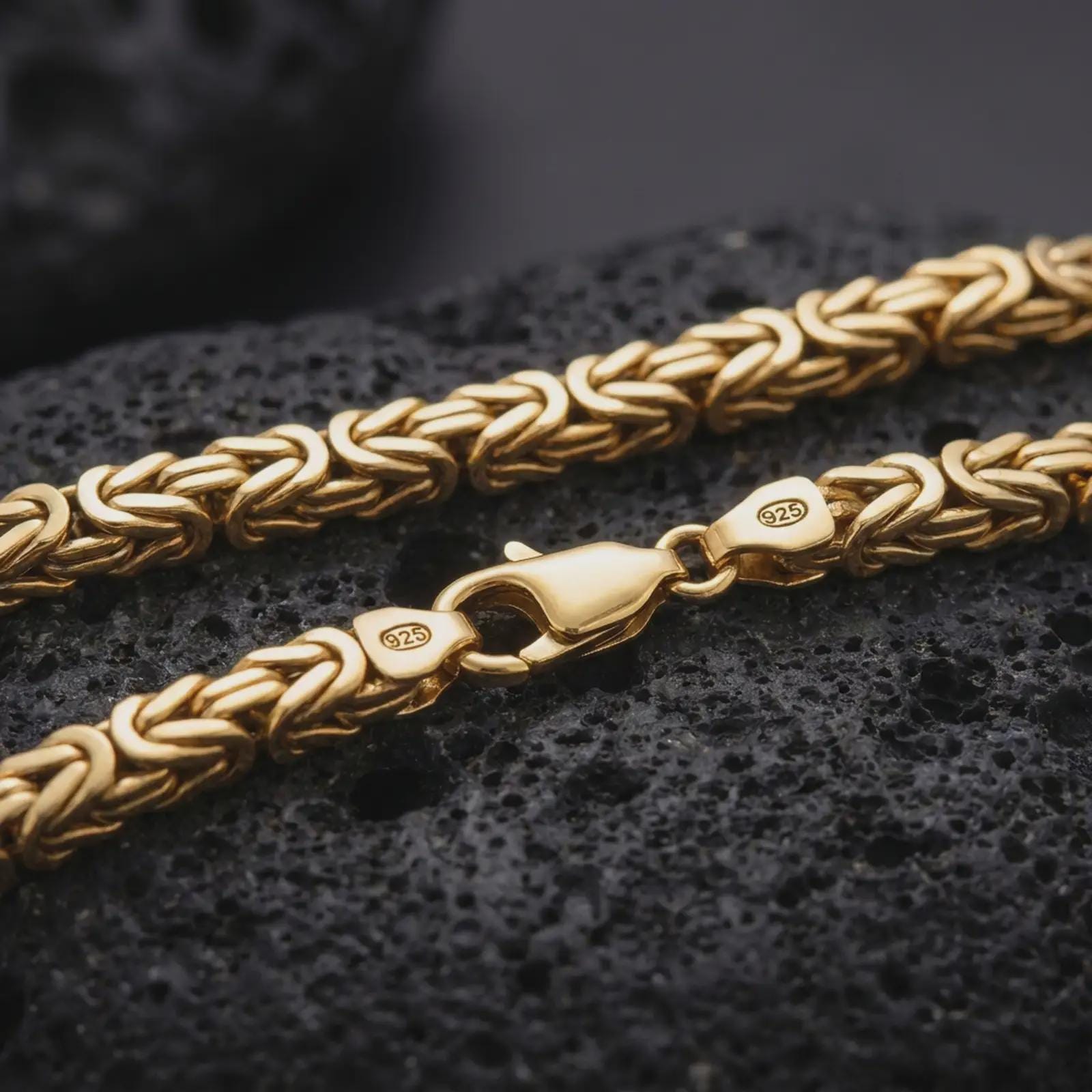 A gold plated Byzantine chain necklace lies across rugged volcanic rock, featuring bold interlocking links, a lobster clasp, and a substantial masculine chain profile in a luxury macro studio close-up.