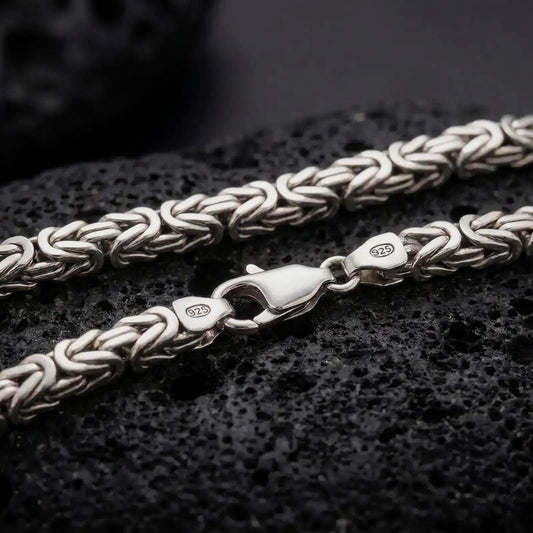 The polished silver Byzantine chain necklace is displayed on textured black volcanic rock, highlighting the rounded woven link pattern, secure clasp, and 925 silver markings in a premium macro photograph.