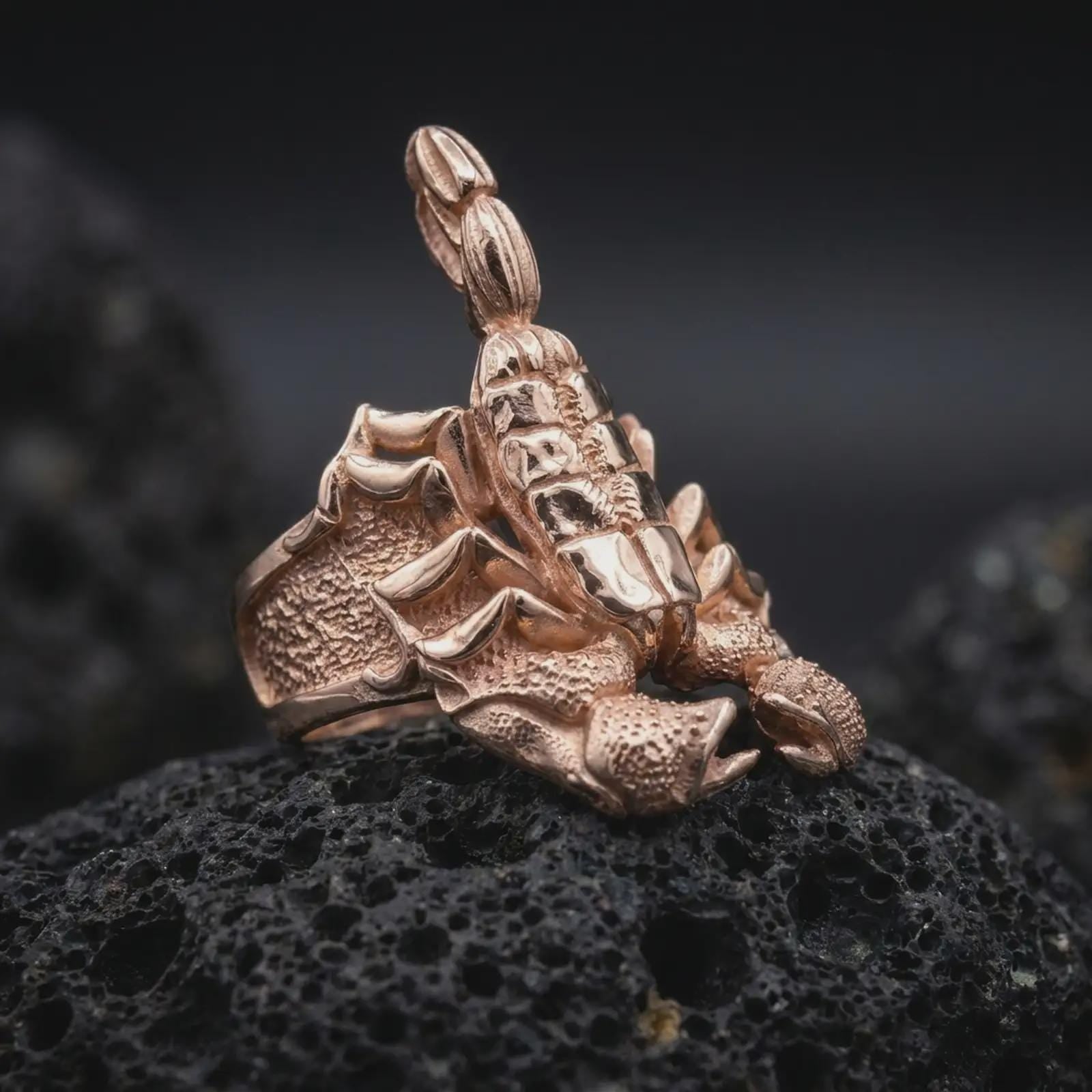 The rose gold plated scorpion ring is arranged on black volcanic rock, emphasizing the wide band, raised scorpion tail, claw details, and gothic animal-inspired construction in a refined macro studio image.