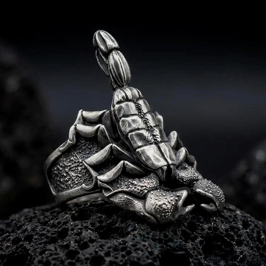 An oxidized sterling silver scorpion ring rests on dark volcanic rock, showing the raised segmented tail, curved stinger, armored body, textured claws, and rugged wide band in a macro studio close-up.