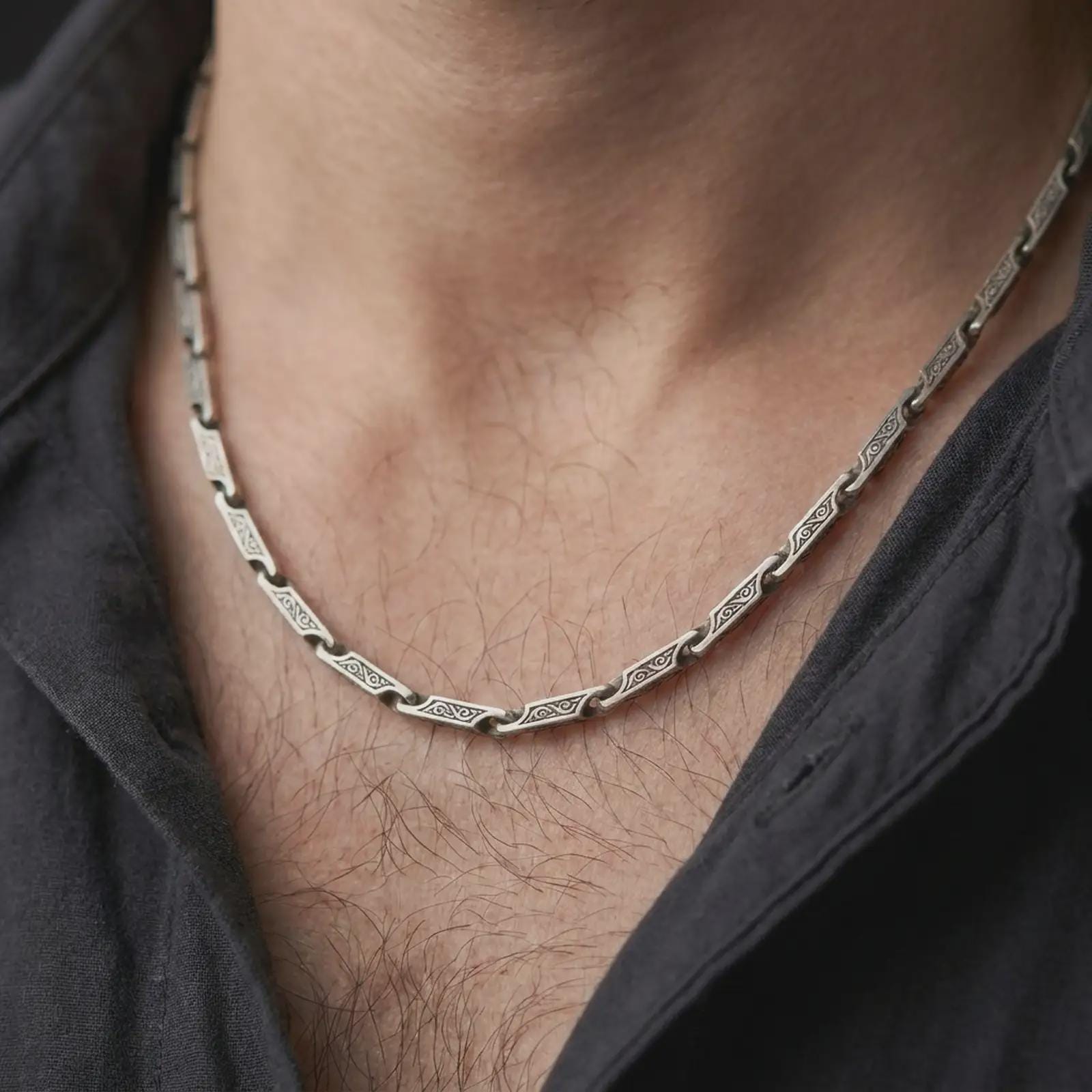 An oxidized scroll chain necklace is worn on a man’s neck, displaying dark recessed engravings, rectangular sterling silver links, and a bold masculine silhouette in a close-up lifestyle shot.