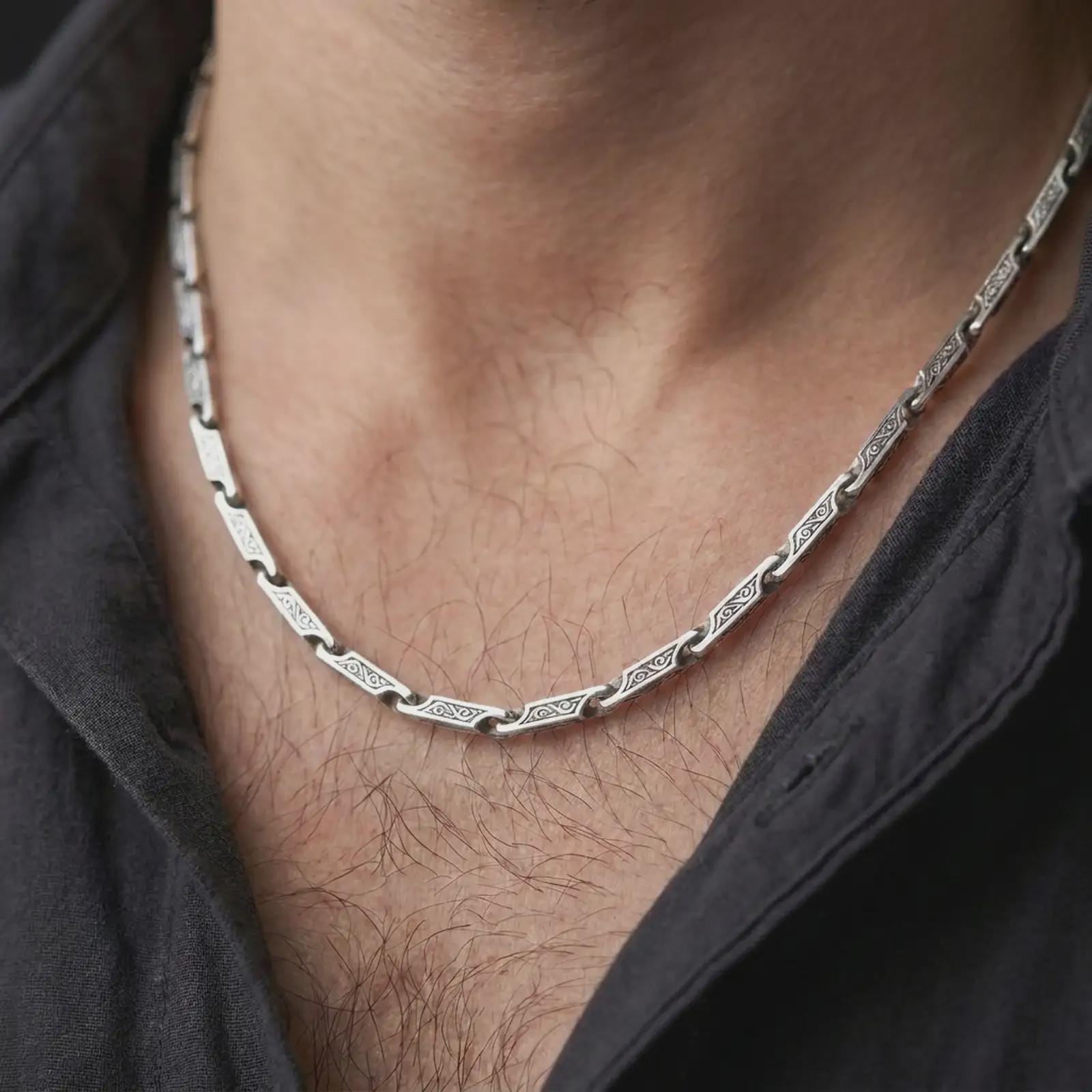 The polished silver ornate bar chain is worn on a man’s chest, showing the repeating engraved link pattern and articulated necklace structure in a premium lifestyle photograph.