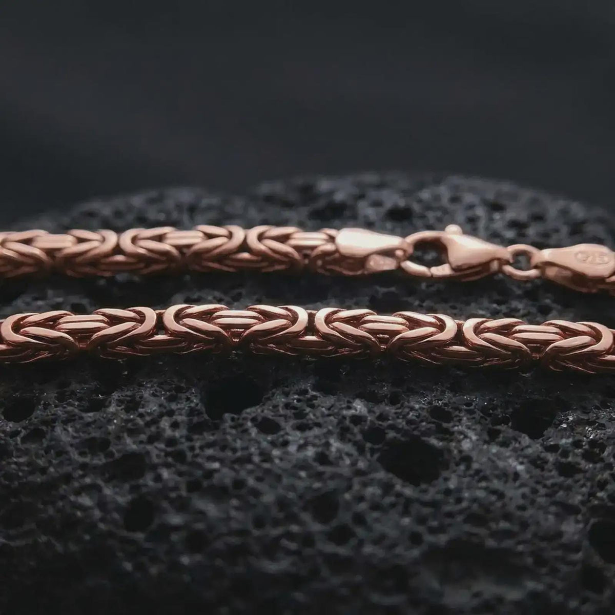 The rose gold plated 4mm square Byzantine chain necklace is arranged on black volcanic rock, featuring a detailed square link structure and elegant woven pattern in a high-end macro image.