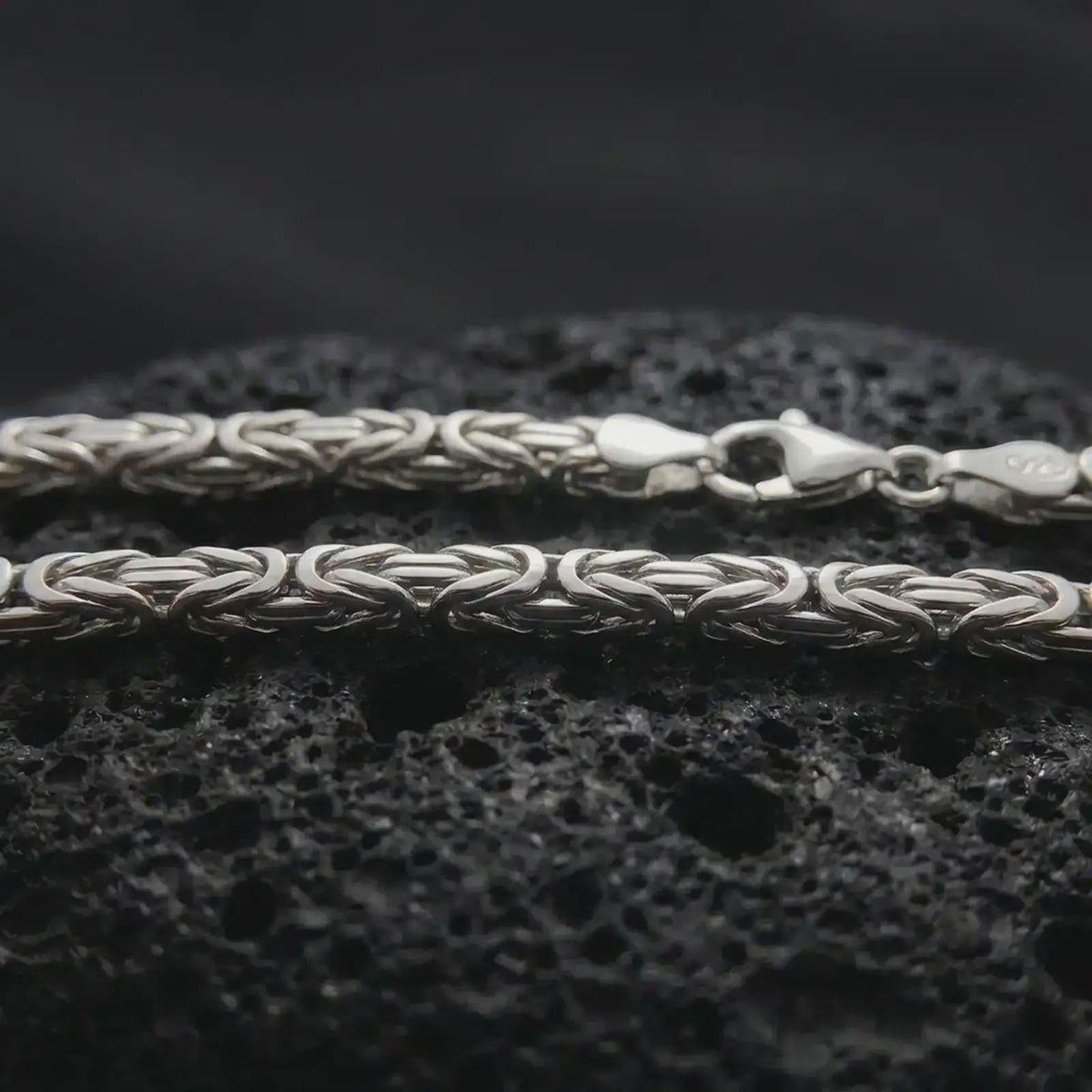 The polished silver 4mm square Byzantine chain necklace is displayed on textured black volcanic rock, emphasizing its intricate link pattern and refined geometric silhouette in a premium macro photograph.