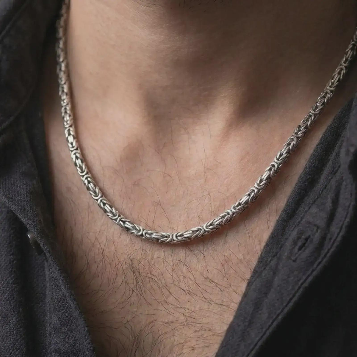 A 5mm square Byzantine chain necklace with an oxidized finish is worn on a man’s neck, displaying dark recessed link details and a strong masculine silhouette in a close-up lifestyle shot.