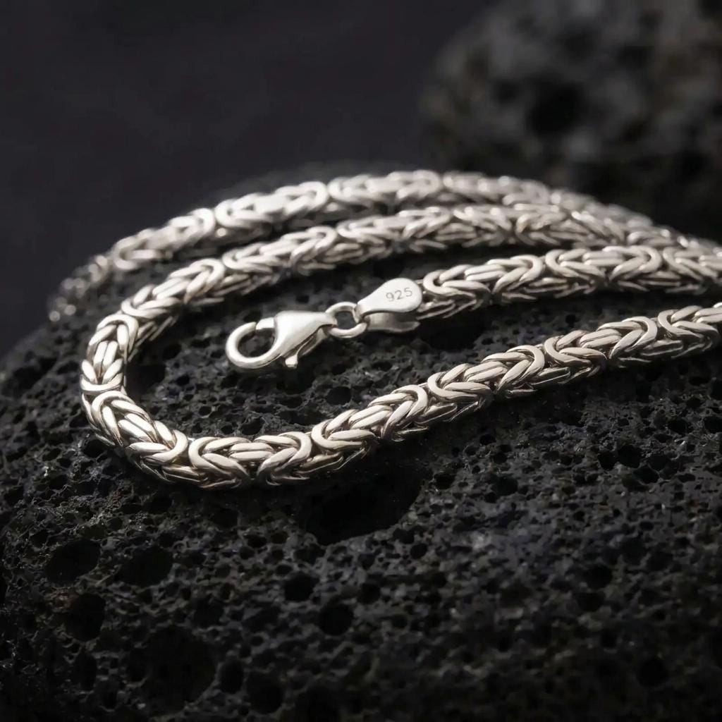 The polished silver 5mm square Byzantine chain necklace is displayed on textured black volcanic rock, highlighting the clean geometric weave, lobster clasp, and 925 silver stamp in a premium macro photograph.