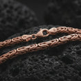 Load image into Gallery viewer, The rose gold plated 4mm Byzantine chain necklace is arranged on black volcanic rock, emphasizing its rounded woven structure, flexible construction, and detailed link pattern in a refined macro image.
