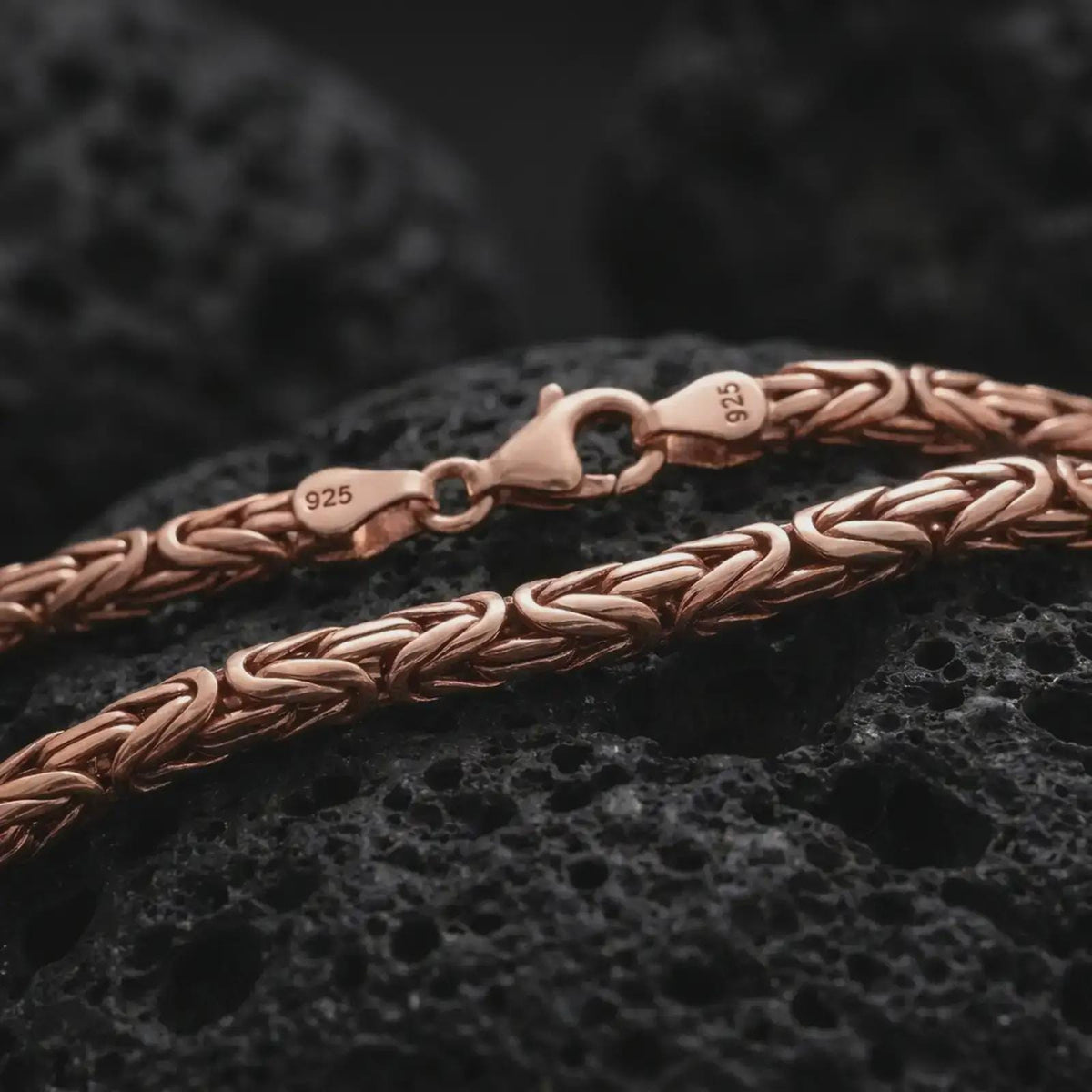 The rose gold plated 4mm Byzantine chain necklace is arranged on black volcanic rock, emphasizing its rounded woven structure, flexible construction, and detailed link pattern in a refined macro image.