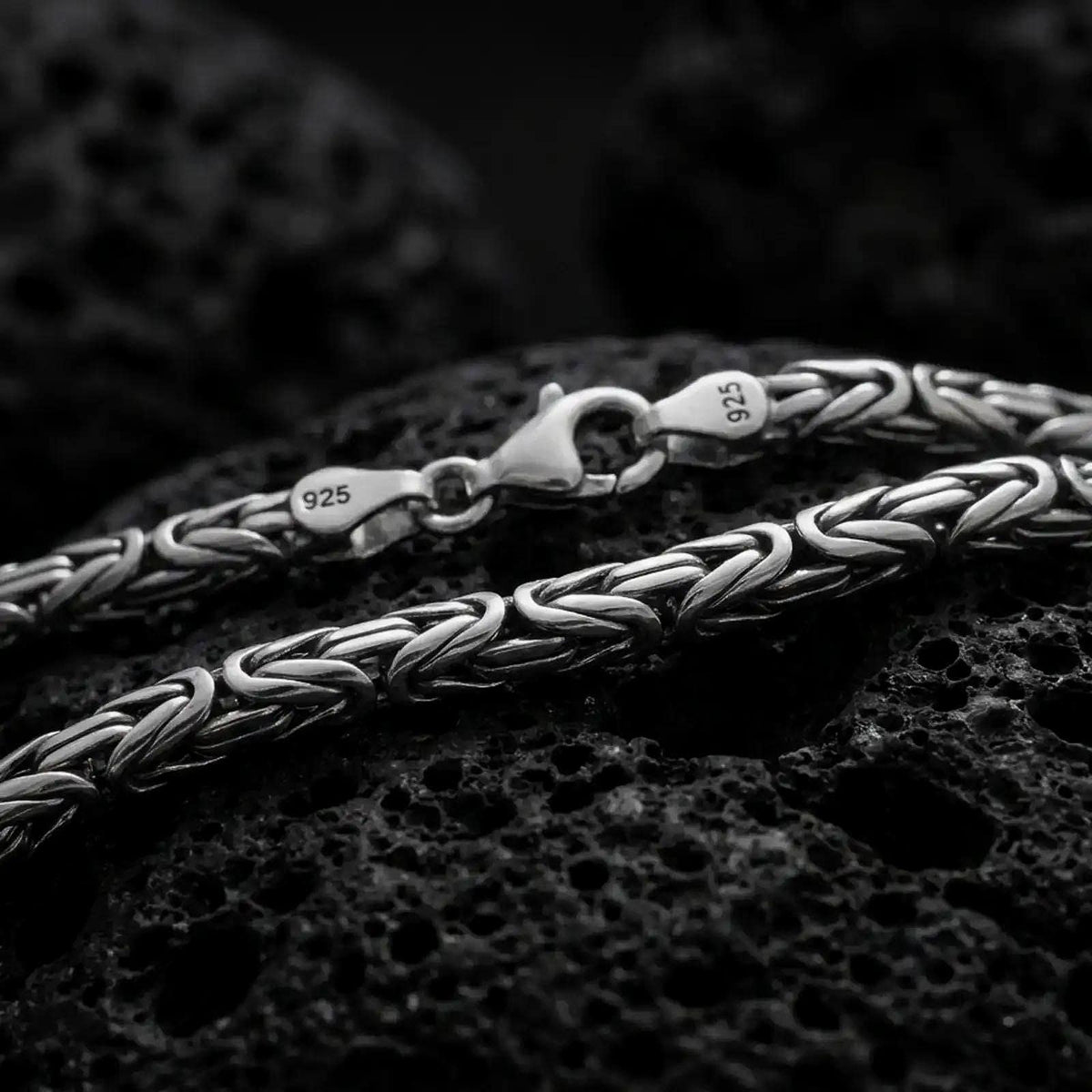 A 4mm round Byzantine chain necklace with an oxidized finish rests on dark volcanic rock, showing its compact interwoven links and dimensional sterling silver texture in a macro studio close-up.