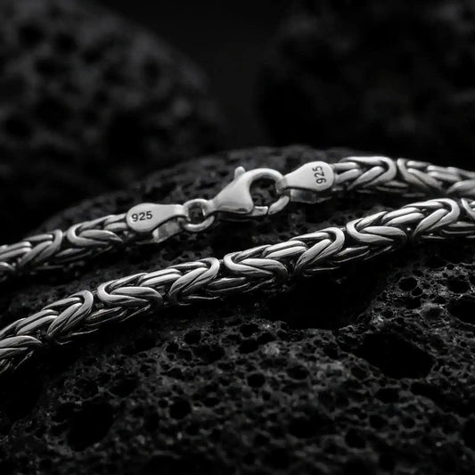 A 4mm round Byzantine chain necklace with an oxidized finish rests on dark volcanic rock, showing its compact interwoven links and dimensional sterling silver texture in a macro studio close-up.