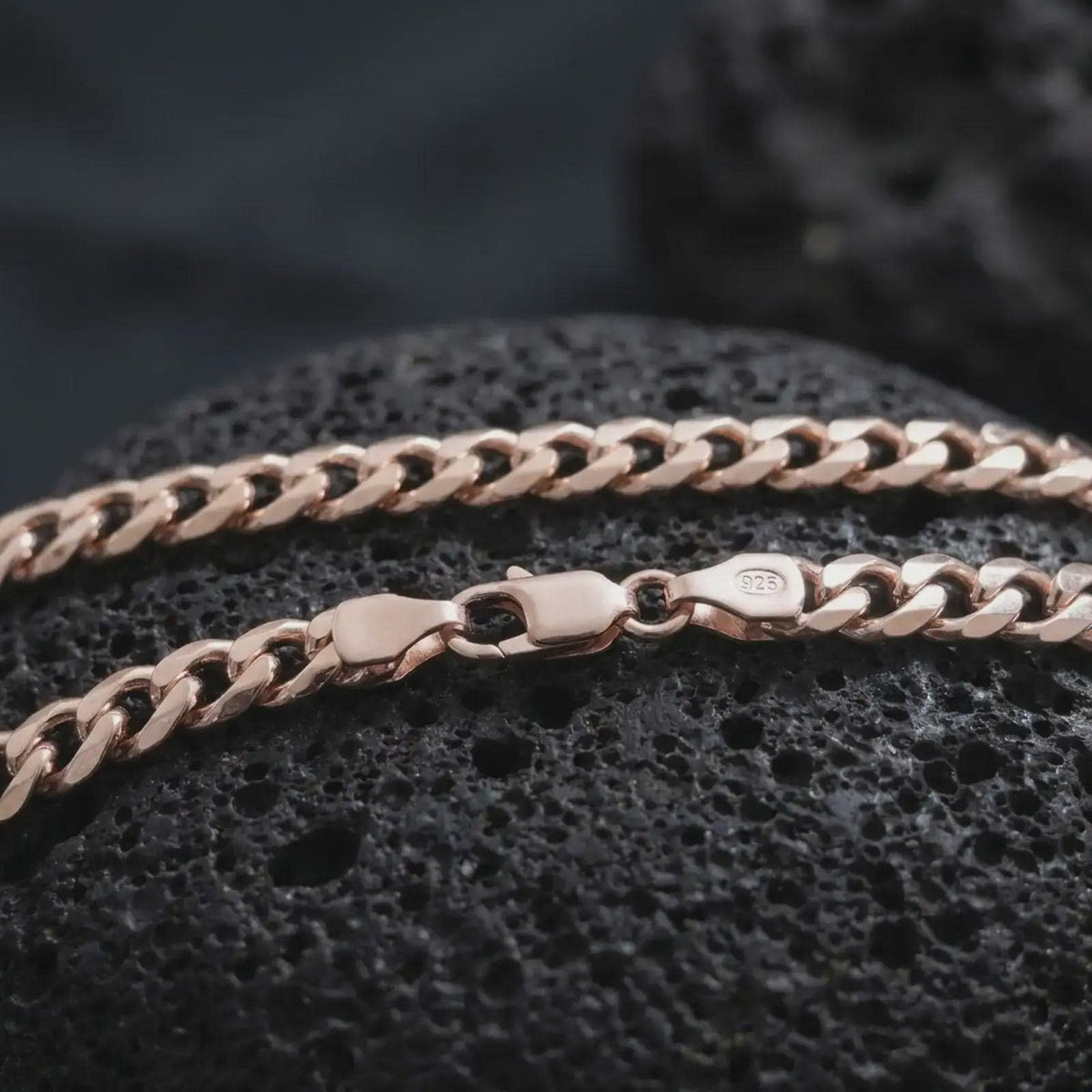 The rose gold plated Cuban chain necklace is arranged on black volcanic rock, emphasizing its 5mm curb link construction, flexible structure, and timeless polished surface in a detailed macro image.
