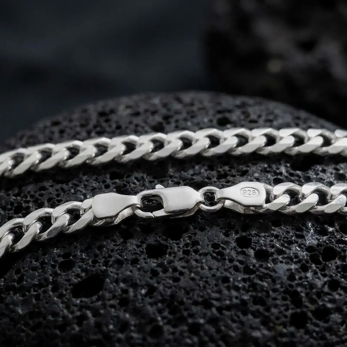 The polished silver 5mm Cuban chain necklace is displayed across textured black volcanic rock, highlighting the interlocking curb link pattern and secure clasp in a premium macro photograph.