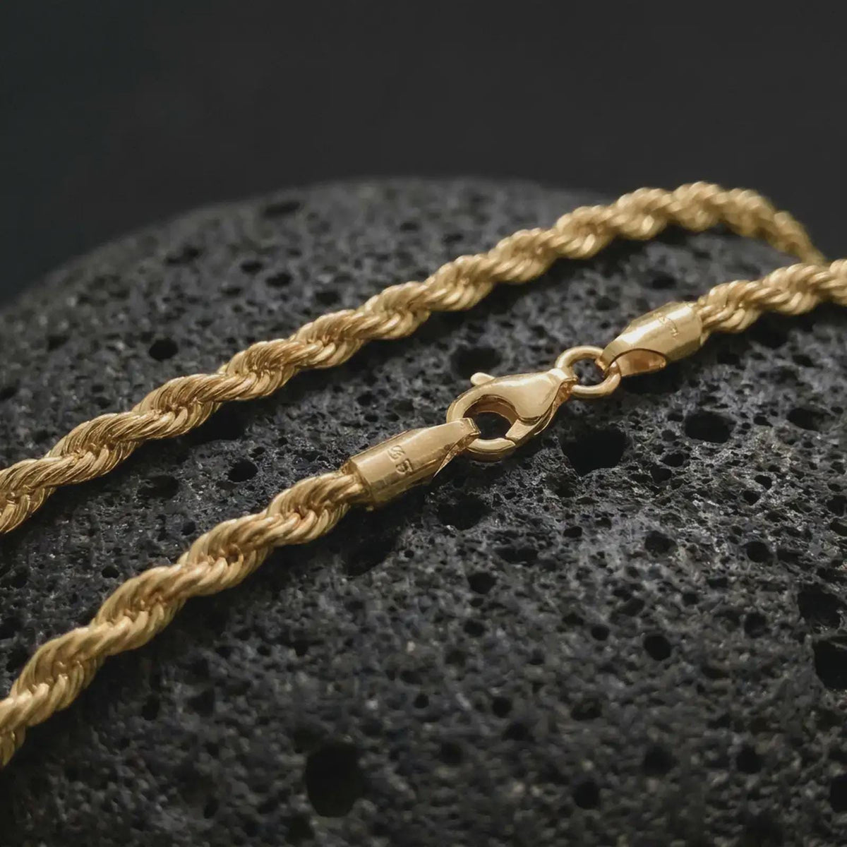 A gold plated 3mm rope chain necklace lies across rugged volcanic rock, featuring a compact twisted link structure and subtle masculine shine in a luxury macro studio close-up.