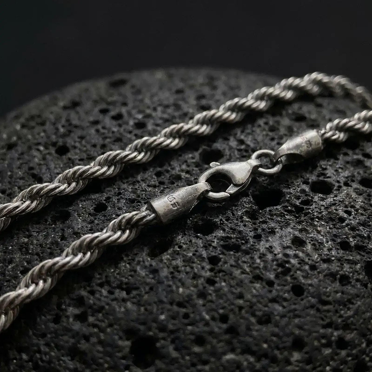 A 3mm rope chain necklace with an oxidized finish rests on dark volcanic rock, showing its tightly twisted sterling silver links and classic spiral texture in a macro studio close-up.